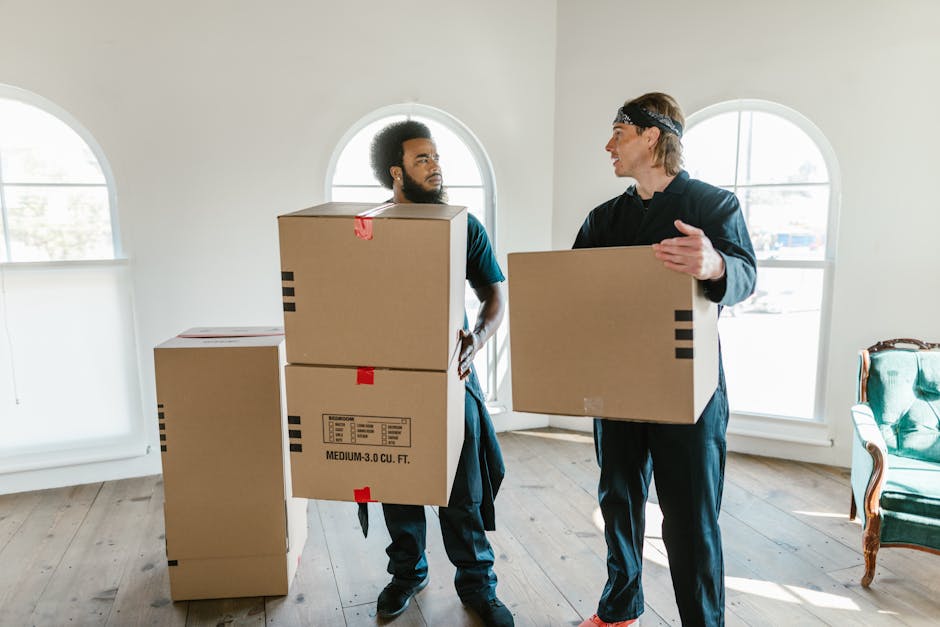A young woman with curly black hair, wearing a light grey top and blue jeans, is sitting on a light wooden floor surrounded by various cardboard boxes of different sizes, some with red and black tape,